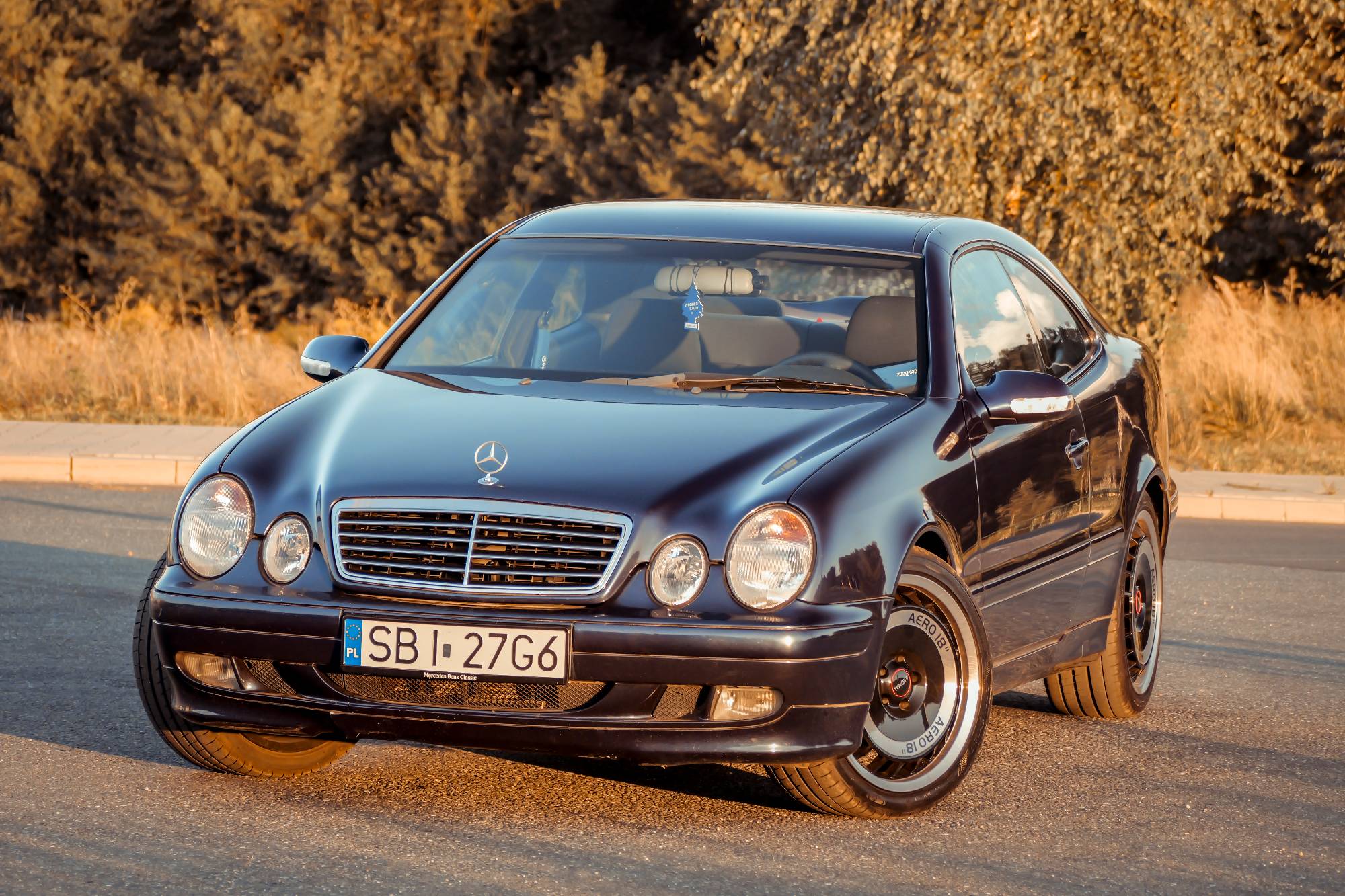 Granatowy Mercedes CLK W208 ujęty z przodu, w tle jesienne drzewa o zachodzie słońca. Fot. Magda (@celty)